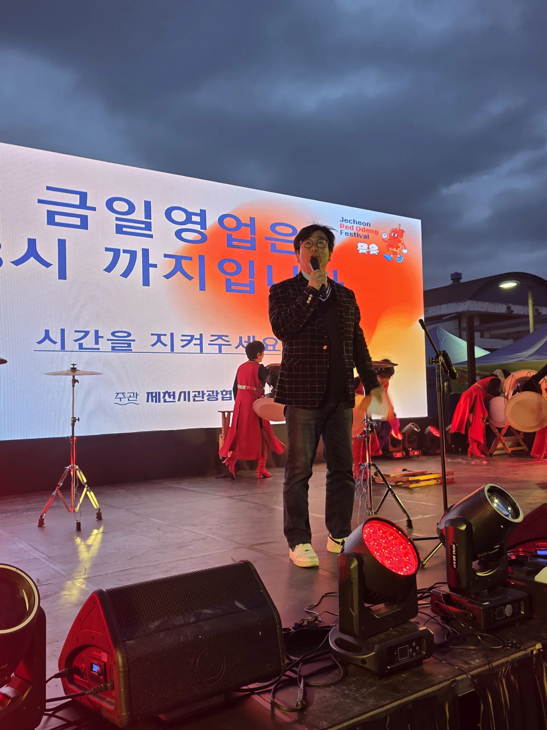 해 질 무렵 야외 무대에서 마이크를 든 채 현수막 앞에 선 MC의 모습