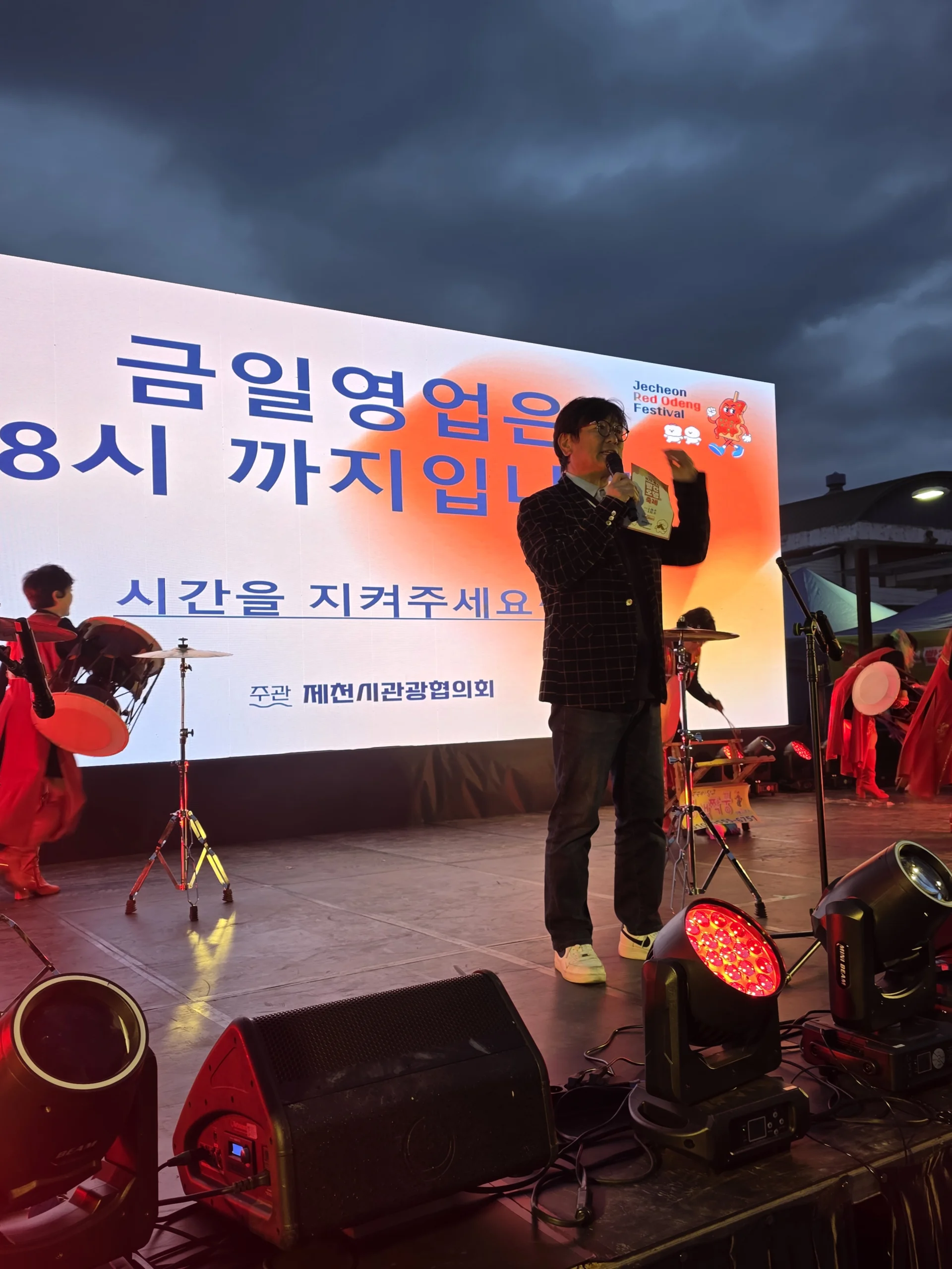 해 질 무렵 야외 무대에서 마이크를 든 채 현수막 앞에 선 MC의 모습