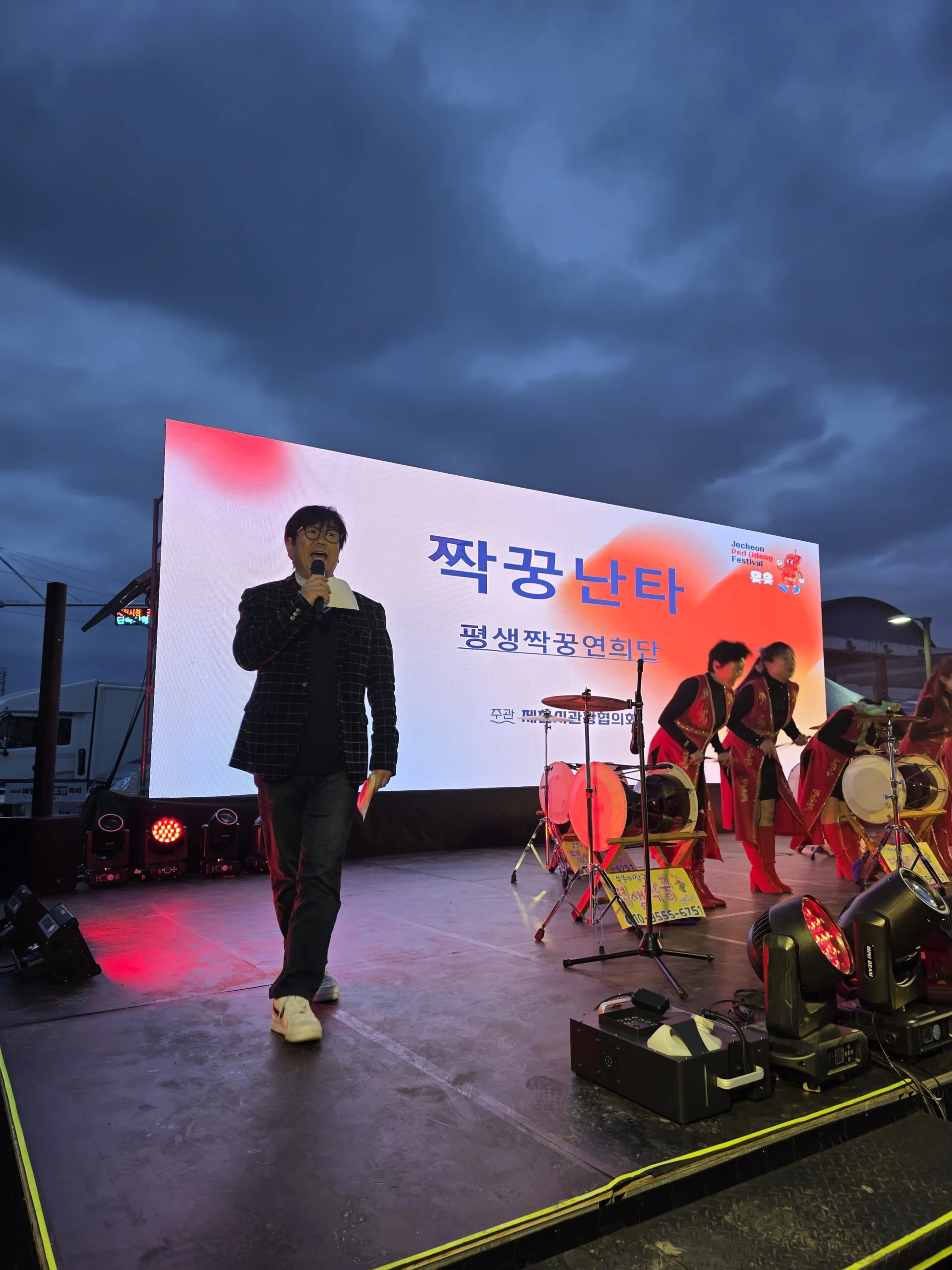 해 질 무렵 야외 무대에 홀로 서서 색색의 배경 앞에서 진행하는 MC의 모습