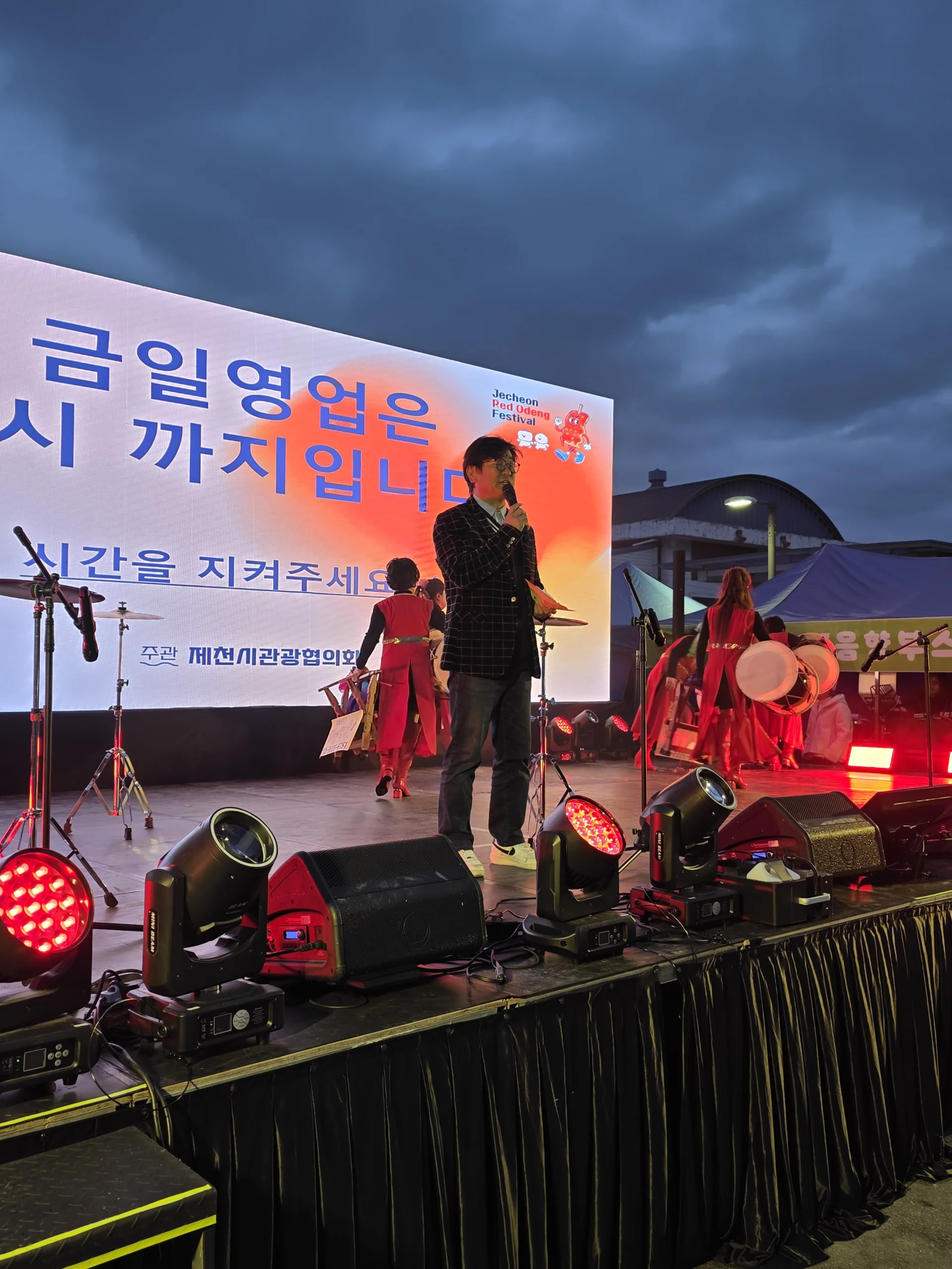 해 질 무렵 야외 무대에서 마이크를 든 채 현수막 앞에 선 MC의 모습