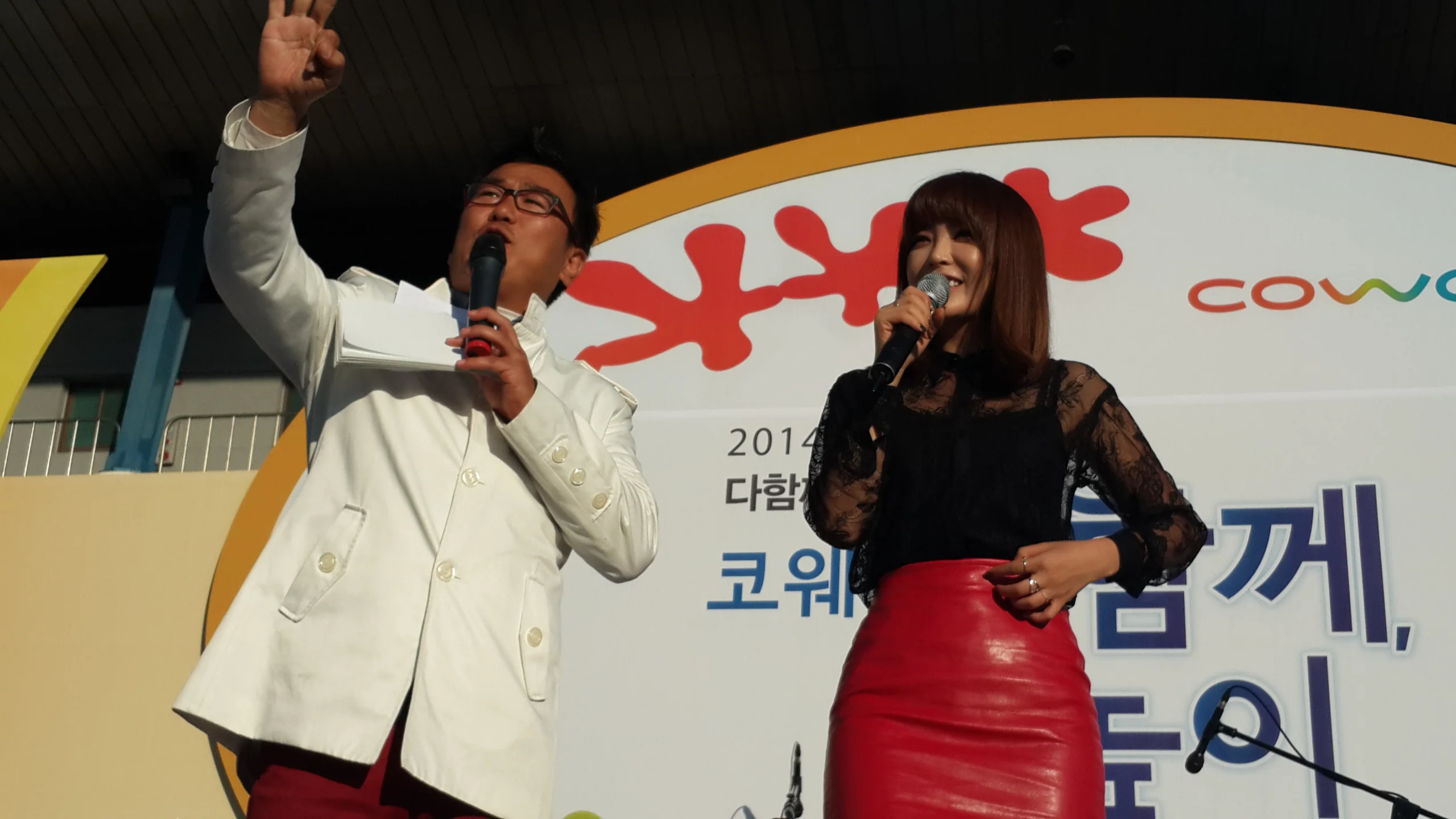 야외 무대에서 컬러풀한 현수막을 배경으로 주먹을 들어올린 사회자