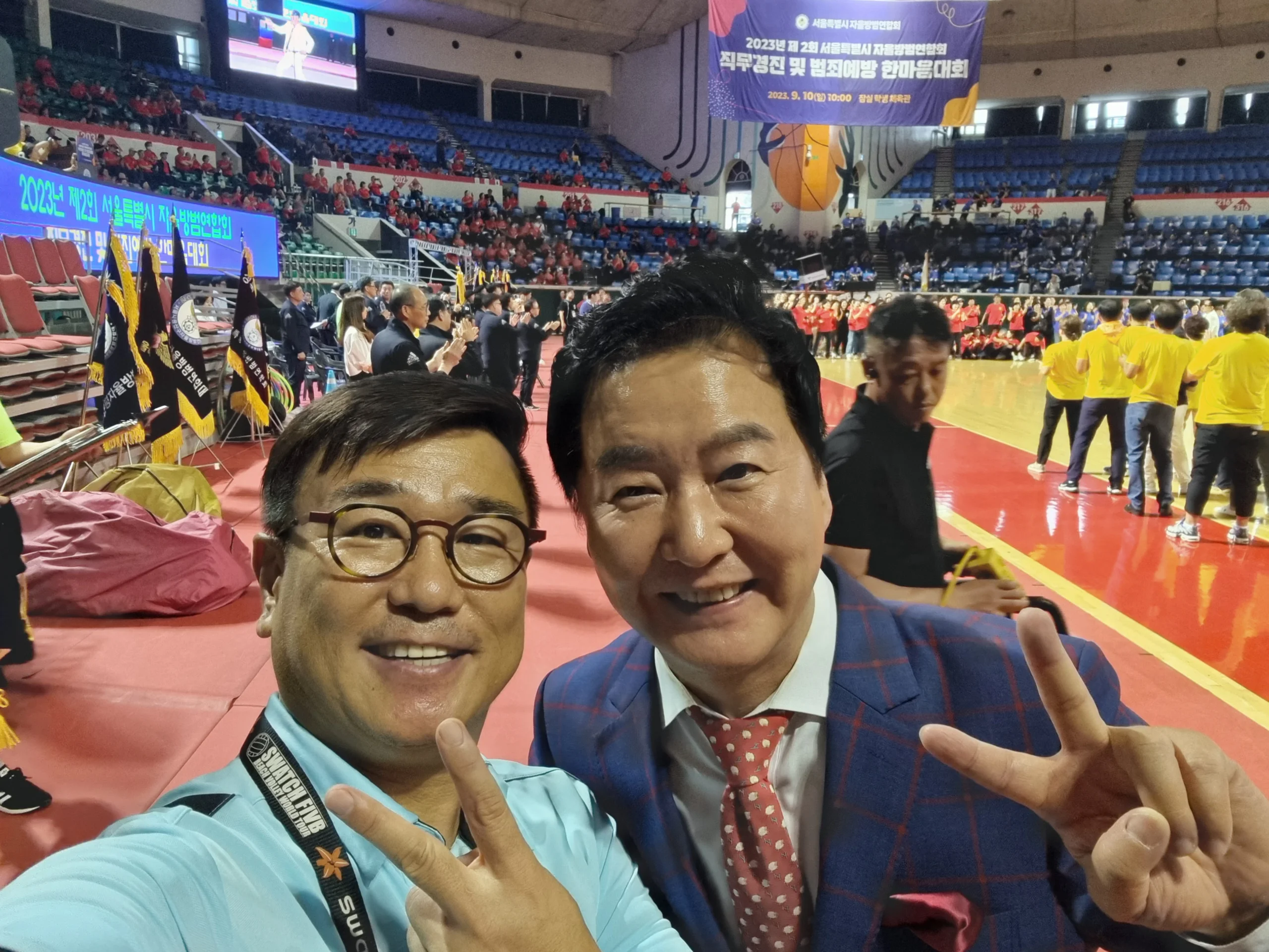 실내 체육관에서 노란색 단체복을 입은 참가자들을 배경으로 포즈를 취한 두 남성