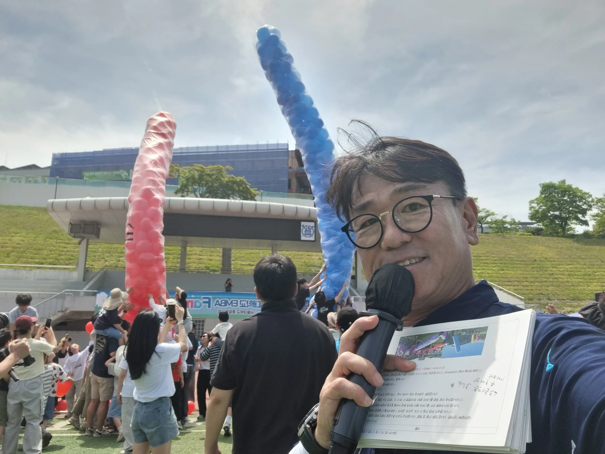 야외에서 마이크와 상패를 든 채 풍선 장식 앞에 선 진행 모습