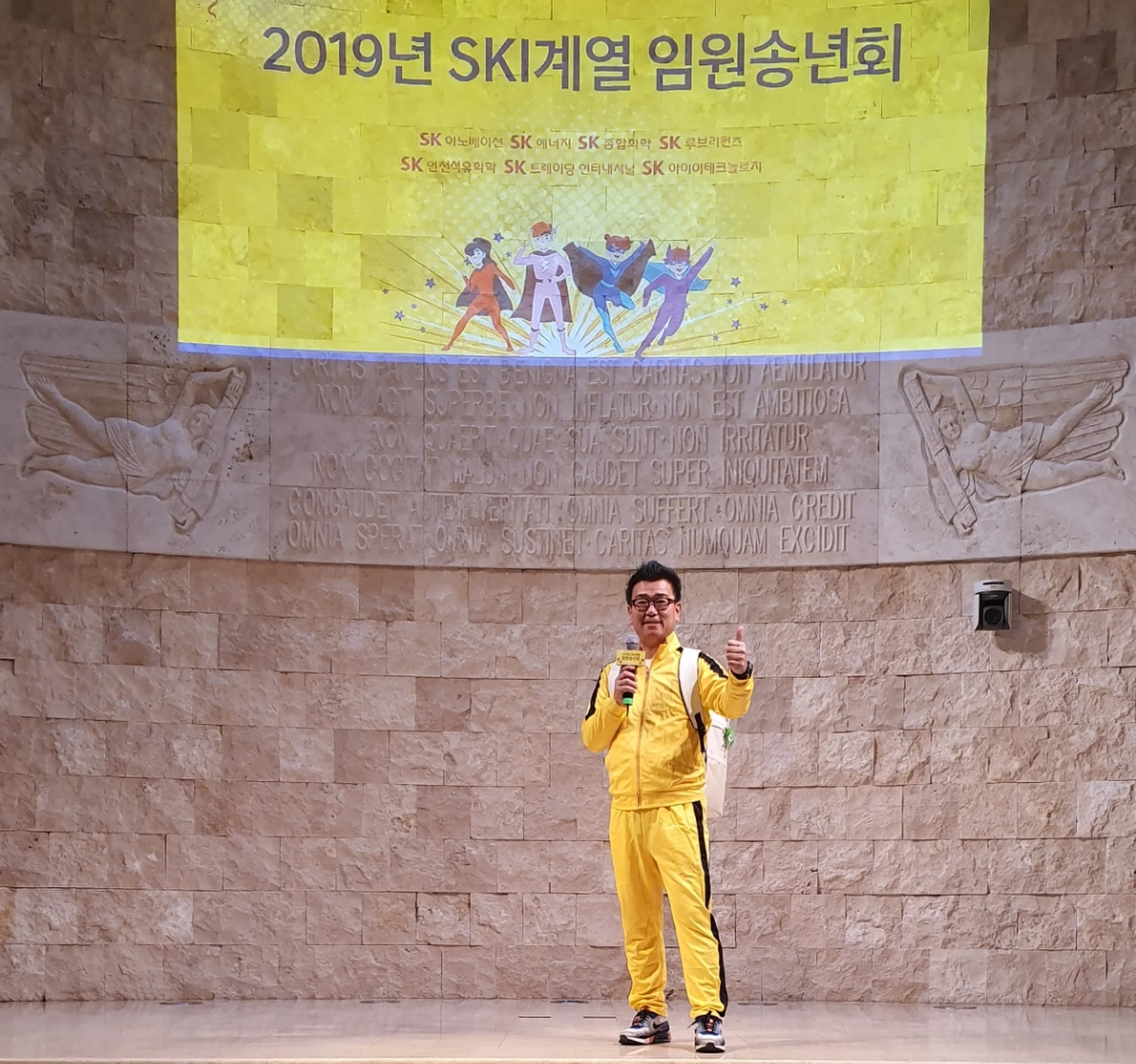 노란색 트레이닝복을 입고 현수막이 걸린 야외 무대에서 춤추는 듯한 동작을 하는 모습