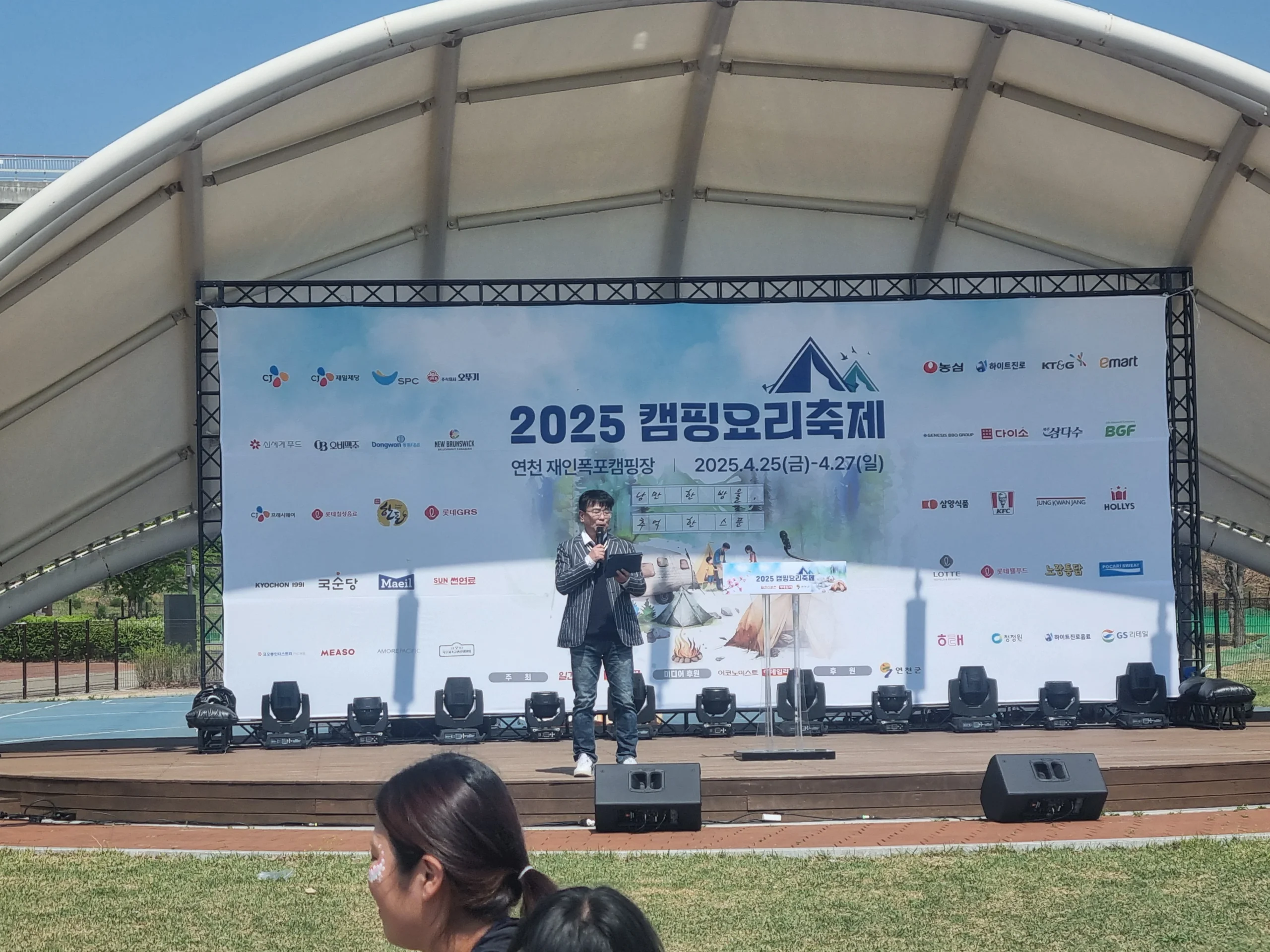 천막을 배경으로 야외 축제 무대에서 마이크를 든 진행자
